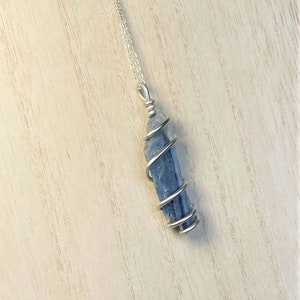 K&ouml;nnte beinhalten: Eine silberne Halskette mit einem Anh&auml;nger aus blauem Kyanitkristall. Der Kristall ist mit silbernem Draht umwickelt und h&auml;ngt an einer feinen Kette. Der Anh&auml;nger hat eine leuchtend blaue Farbe, und die Halskette wird auf einer hellen Holzoberfl&auml;che pr&auml;sentiert.