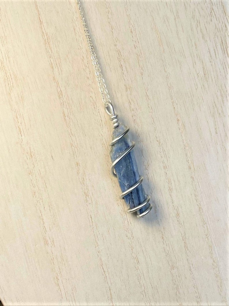 K&ouml;nnte beinhalten: Eine silberne Halskette mit einem Anh&auml;nger aus blauem Kyanitkristall. Der Kristall ist mit silbernem Draht umwickelt und h&auml;ngt an einer feinen Kette. Der Anh&auml;nger hat eine leuchtend blaue Farbe, und die Halskette wird auf einer hellen Holzoberfl&auml;che pr&auml;sentiert.