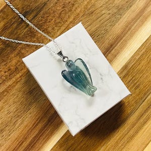 May include: A silver necklace with a pendant shaped like an angel. The pendant is a translucent blue-green color and is resting on a white box with a marble pattern. The background is a wooden surface.