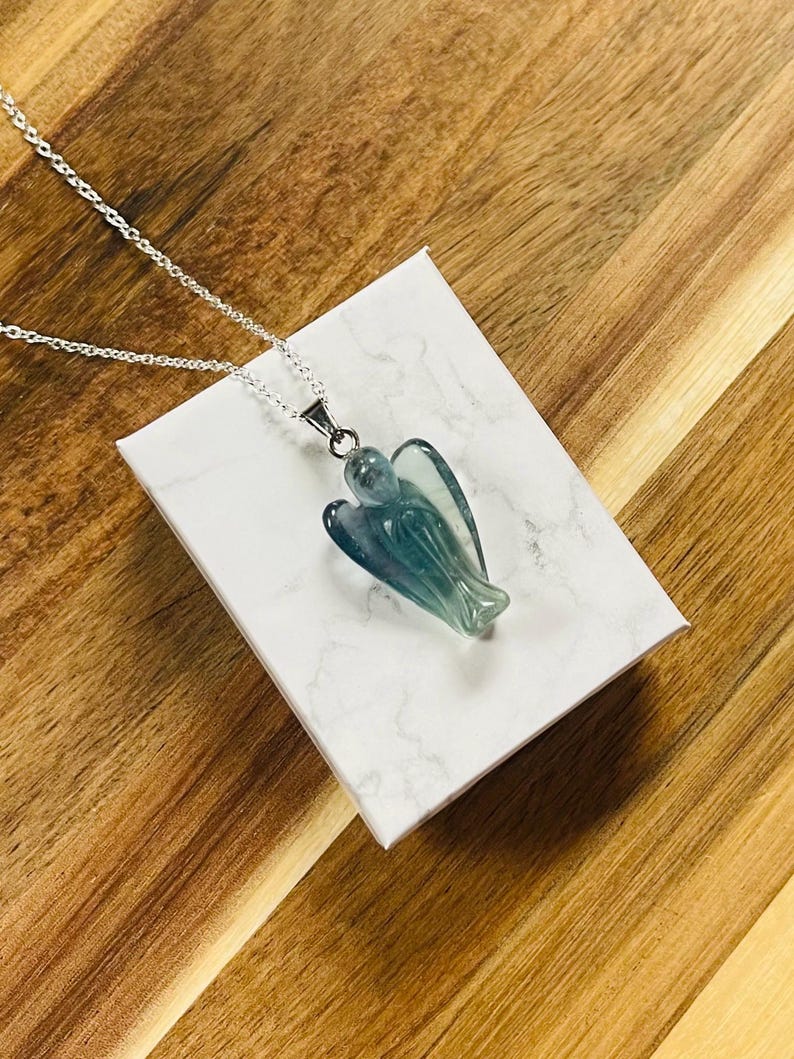 May include: A silver necklace with a pendant shaped like an angel. The pendant is a translucent blue-green color and is resting on a white box with a marble pattern. The background is a wooden surface.