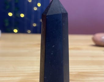 Può includere: Un alto obelisco di cristallo nero è posto su una superficie di legno. Il cristallo ha un design sfaccettato e una parte superiore appuntita. Lo sfondo presenta un'esposizione sfocata di luci e un muro viola.