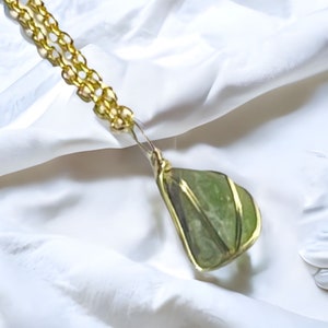 Puede incluir: Un collar de cadena de oro con un colgante de piedra preciosa verde. La piedra preciosa está envuelta en alambre de oro y tiene una forma facetada.
