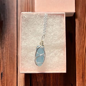 May include: A silver chain necklace with a light blue sea glass pendant wrapped in silver wire. The necklace is displayed in a white gift box with a light blue lining.