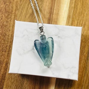 May include: A silver chain necklace with a pendant carved in the shape of an angel. The angel pendant is a translucent blue-green color. The necklace is displayed on a white box with a marble pattern.