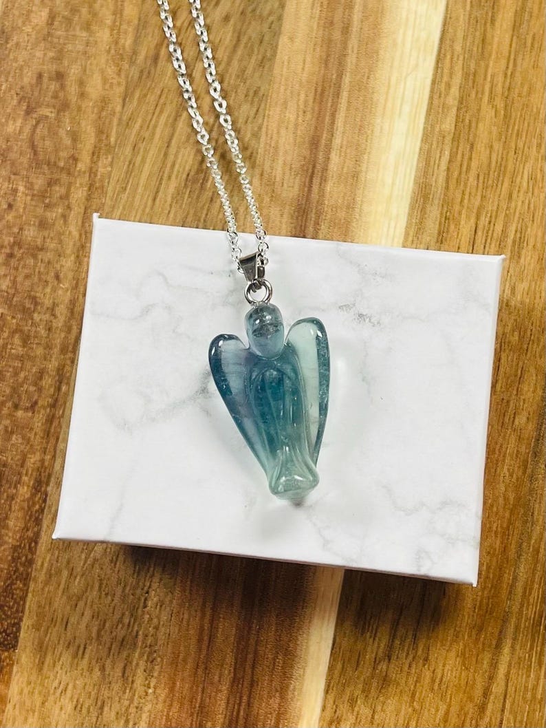 May include: A silver chain necklace with a pendant carved in the shape of an angel. The angel pendant is a translucent blue-green color. The necklace is displayed on a white box with a marble pattern.