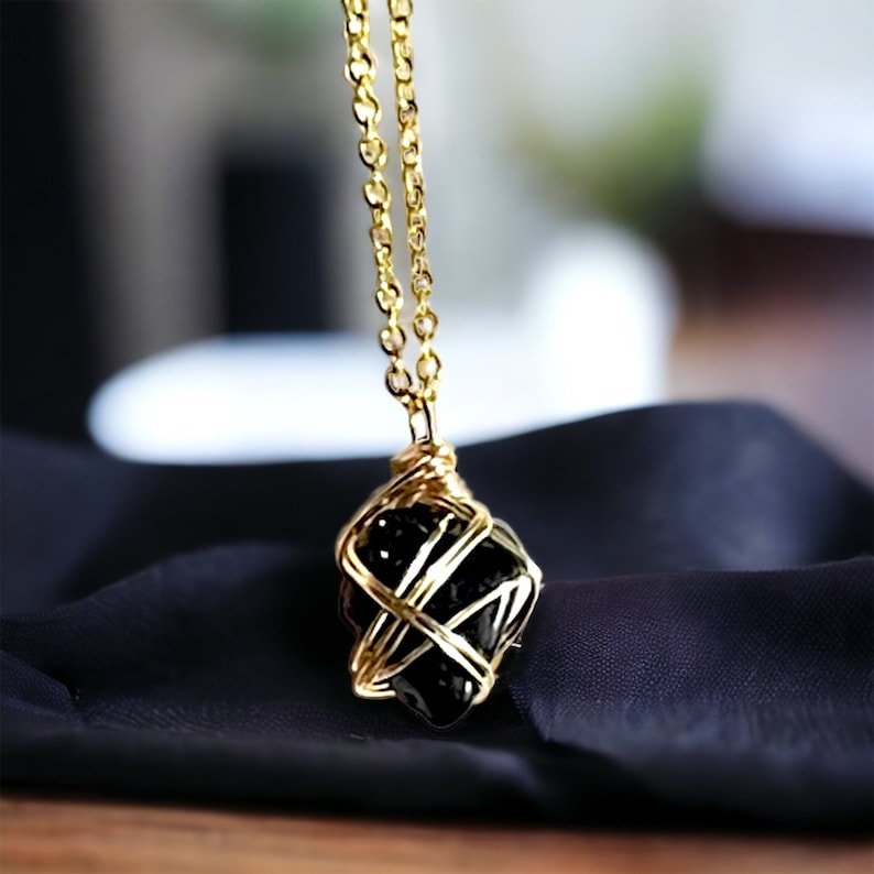 May include: A black gemstone pendant wrapped in gold wire, hanging from a gold chain necklace. The pendant is on a black fabric background.