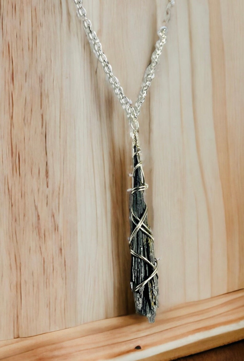 May include: A black kyanite pendant necklace with a silver-colored chain. The kyanite is wrapped in silver-colored wire. The necklace is displayed against a light brown wooden background.
