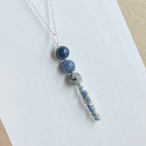 Peut inclure: Un collier en chaîne argent avec un pendentif en pierre bleue et blanche. Le pendentif présente trois pierres bleues rondes et une longue pierre bleue mince enveloppée de fil d'argent.