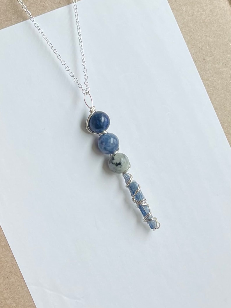 May include: A silver chain necklace with a blue and white stone pendant. The pendant features three round blue stones and a long, thin blue stone wrapped in silver wire.