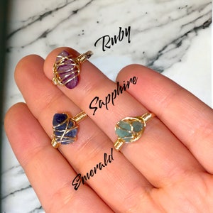 Puede incluir: Tres anillos de alambre dorado envueltos con piedras preciosas de diferentes colores. Los anillos están etiquetados como "Ruby", "Sapphire" y "Emerald".