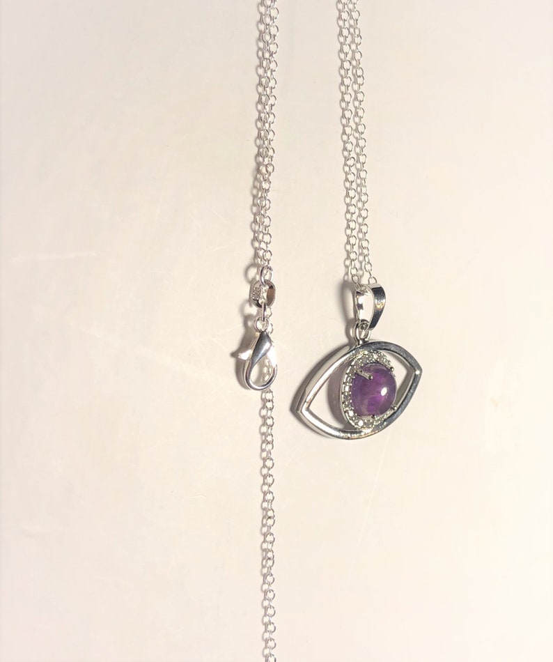Op de afbeelding: Een zilveren ketting met een oogvormige hanger met een centrale ovale paarse cabochonsteen, omringd door een halo van kleine heldere steentjes. De hanger hangt aan een fijne zilveren ketting die is voorzien van een karabijnsluiting.