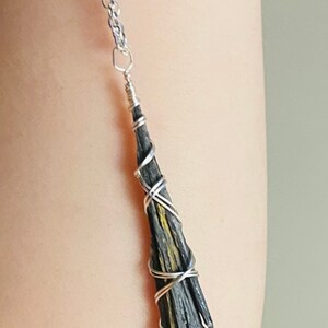 May include: A silver chain necklace with a black and yellow kyanite pendant. The kyanite is wrapped in silver wire, creating a unique and naturalistic design. The pendant is approximately 2 inches long.