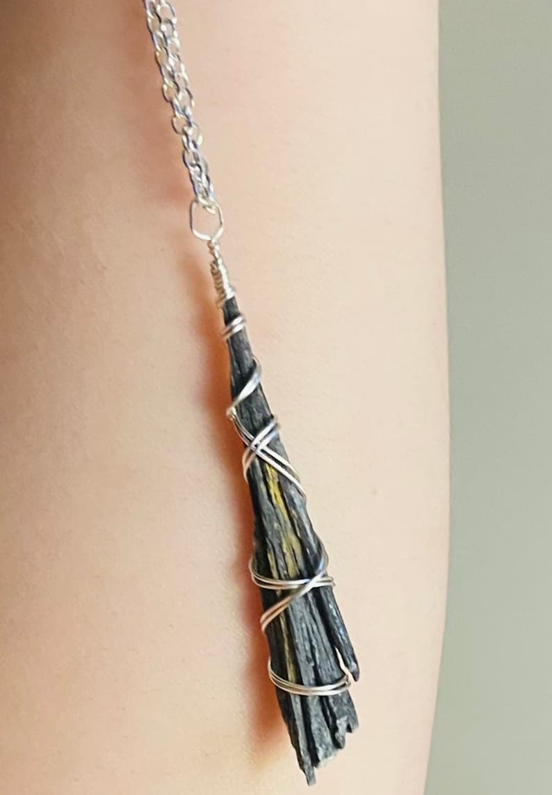 May include: A silver chain necklace with a black and yellow kyanite pendant. The kyanite is wrapped in silver wire, creating a unique and naturalistic design. The pendant is approximately 2 inches long.