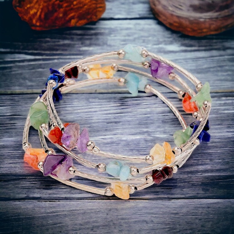 Op de afbeelding: Een meerstrengs wikkelarmband met zilverkleurig metaaldraad en kleine, onregelmatige, gepolijste edelsteensplinters in diverse kleuren zoals paars, blauw, groen, oranje en geel, afgewisseld met kleine zilverkleurige metalen kralen.