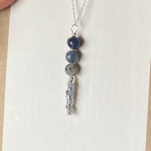 May include: A silver chain necklace with a blue kyanite crystal pendant. The pendant features three round blue kyanite beads and a long, thin blue kyanite crystal wrapped in silver wire.