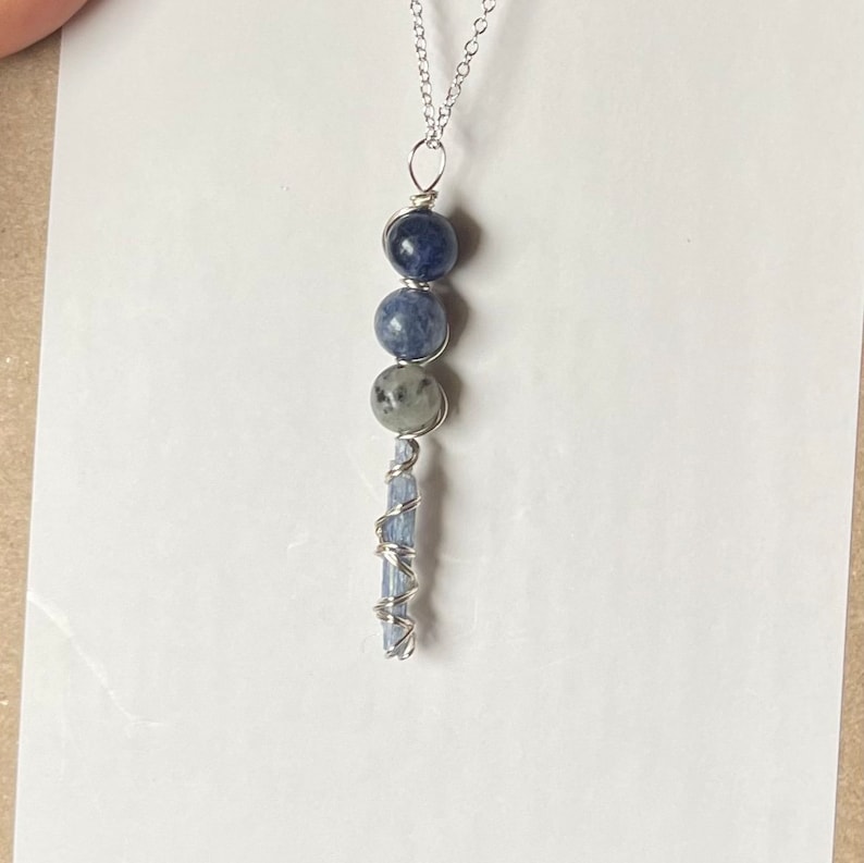 May include: A silver chain necklace with a blue kyanite crystal pendant. The pendant features three round blue kyanite beads and a long, thin blue kyanite crystal wrapped in silver wire.