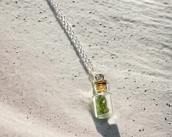 Op de afbeelding: Een zilveren ketting met een klein glazen flesje als hanger, gevuld met groene kristallen. Het flesje heeft een kurken stop.