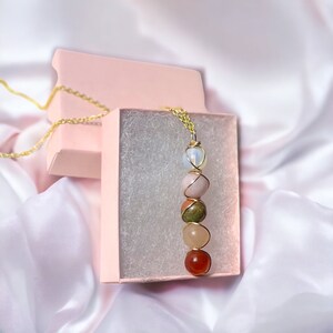 May include: A gold-colored chain necklace with a pendant featuring a vertical arrangement of colorful gemstones. The gemstones include white, pink, green, peach, and red-orange stones, all wire-wrapped. The necklace is presented in a pink gift box.
