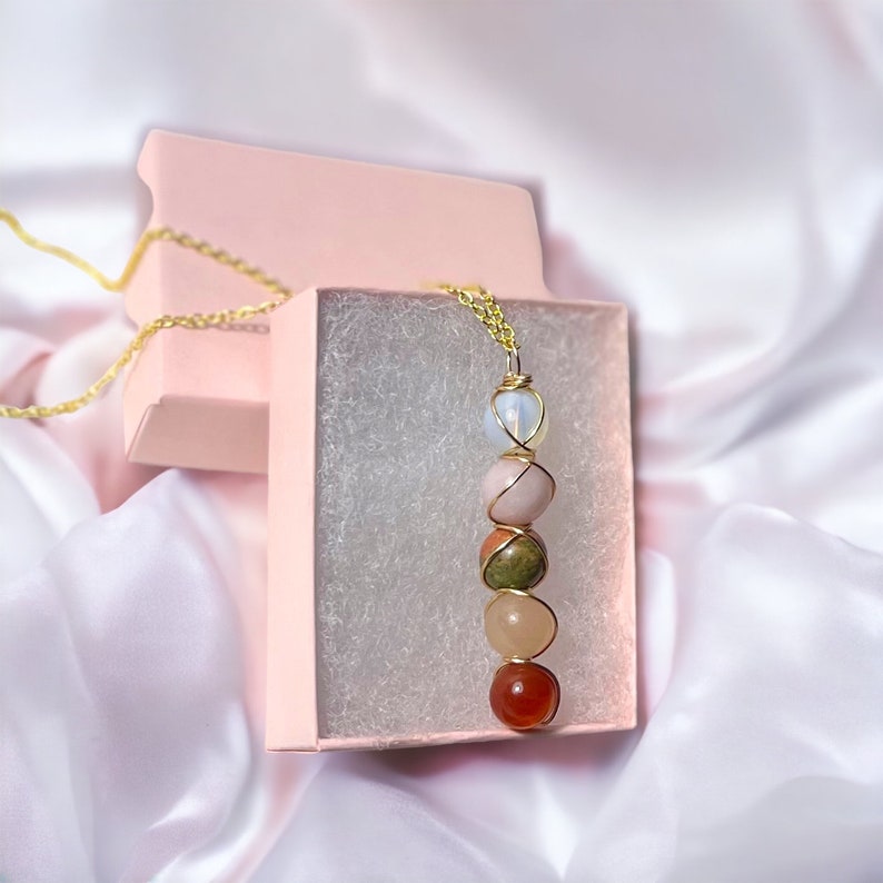 May include: A gold-colored chain necklace with a pendant featuring a vertical arrangement of colorful gemstones. The gemstones include white, pink, green, peach, and red-orange stones, all wire-wrapped. The necklace is presented in a pink gift box.