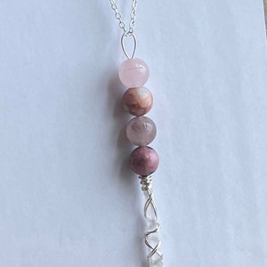 May include: A silver chain necklace with a pink and white crystal pendant. The pendant features a clear crystal wrapped in silver wire and three pink and white gemstone beads.
