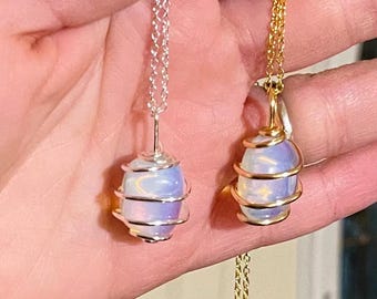 May include: Two necklaces with opalescent, oval-shaped stones. One necklace has a silver chain and wire wrap, the other has a gold chain and wire wrap. The stones have a soft, iridescent glow, with hints of blue and purple.