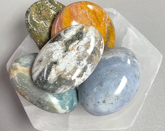 May include: A selection of polished stones in shades of green, orange, blue, and grey, displayed on a white hexagonal dish. The stones have a smooth, glossy surface, and the dish offers a clean, minimalist background.