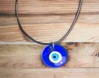 Pode incluir: Um colar com um amuleto de olho turco azul em um cordão preto. O amuleto é redondo e tem um olho branco com uma pupila preta.