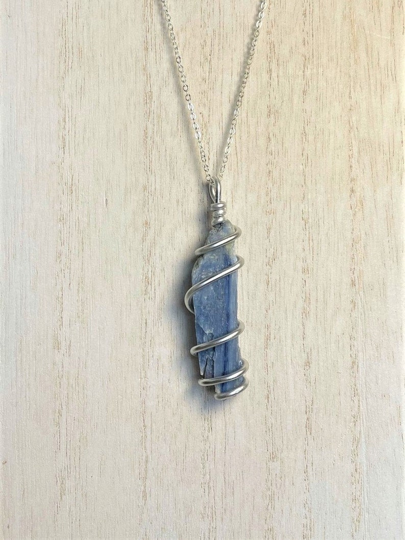 K&ouml;nnte beinhalten: Ein blauer Kyanit-Kristallanh&auml;nger, umwickelt mit silberfarbenem Draht, h&auml;ngt an einer filigranen Silberkette. Der Kristall hat eine leuchtend blaue Farbe und ein nat&uuml;rliches, strukturiertes Aussehen. Die Halskette wird vor einem hellen Holzmaserungshintergrund pr&auml;sentiert.
