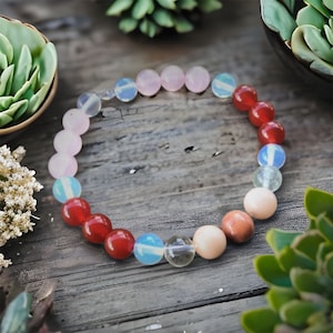 Op de afbeelding: Een kralenarmband met een cirkelvormige opstelling van gladde, gepolijste, bolvormige kralen in diverse kleuren, waaronder doorschijnend lichtblauw, ondoorzichtig lichtroze, dieprood, transparant en zachte perziktinten in een lus.