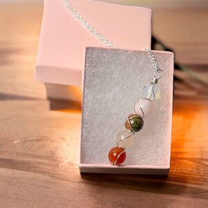 May include: A gemstone necklace with a silver chain and wire wrapping. The necklace features a series of colorful, round stones, including pink, white, orange, and green. The necklace is presented in a pink gift box with a white interior.