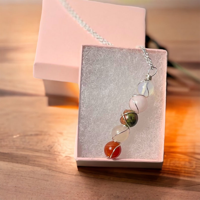 May include: A gemstone necklace with a silver chain and wire wrapping. The necklace features a series of colorful, round stones, including pink, white, orange, and green. The necklace is presented in a pink gift box with a white interior.