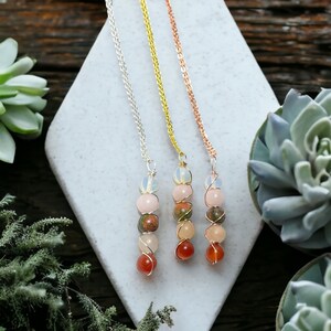 May include: Three gemstone necklaces with silver, gold, and rose gold chains. Each necklace features a series of colorful, round gemstones, including pink, orange, and green stones, wire-wrapped together. The necklaces are displayed on a white, geometric surface.
