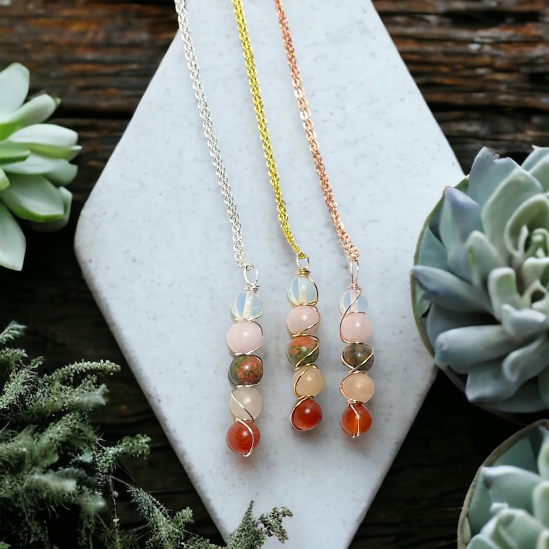 May include: Three gemstone necklaces with silver, gold, and rose gold chains. Each necklace features a series of colorful, round gemstones, including pink, orange, and green stones, wire-wrapped together. The necklaces are displayed on a white, geometric surface.