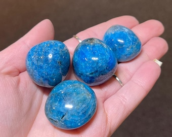 May include: Four polished, oval-shaped blue apatite stones. The stones vary slightly in shade, with some showing darker blue hues and subtle internal patterns. The smooth, rounded forms are held in a hand, showcasing their natural beauty.
