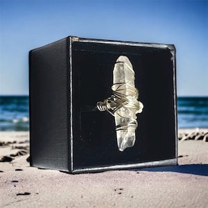 May include: A clear quartz crystal wrapped in gold wire, displayed in a black box. The box is sitting on a sandy beach.