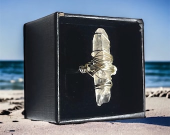 May include: A clear quartz crystal wrapped in gold wire, displayed in a black box. The box is sitting on a sandy beach.