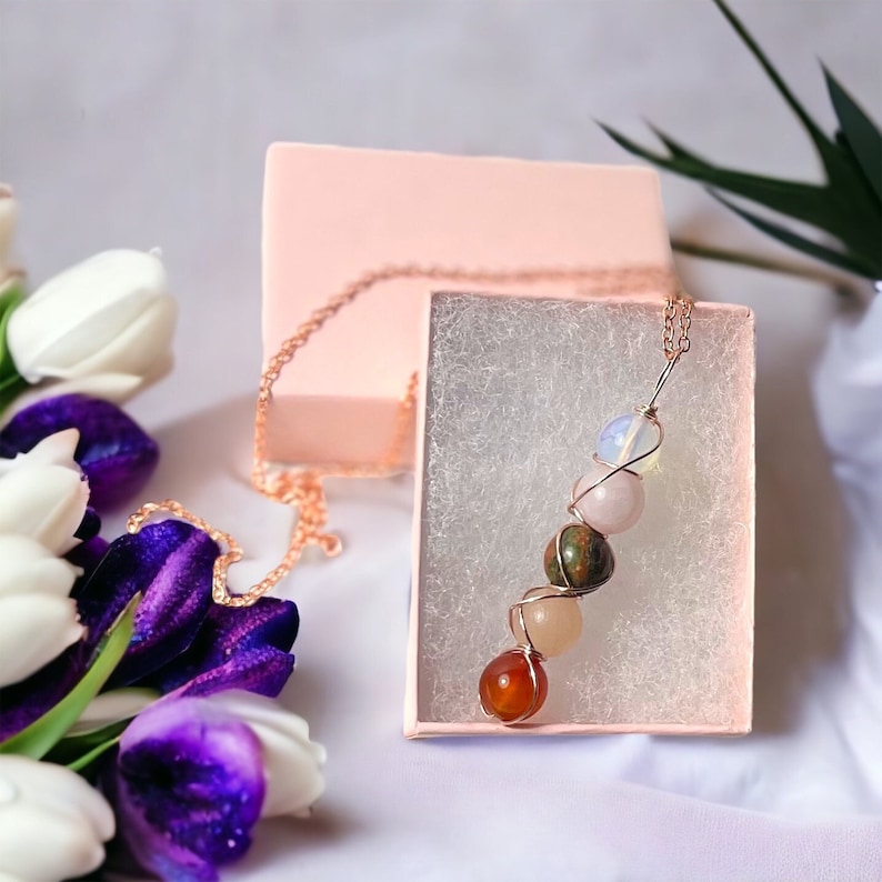 May include: A copper-colored chain necklace with a pendant of various colorful gemstones, including orange, pink, green, and white. The necklace is displayed in a pink gift box with a white interior. White and purple tulips are in the background.