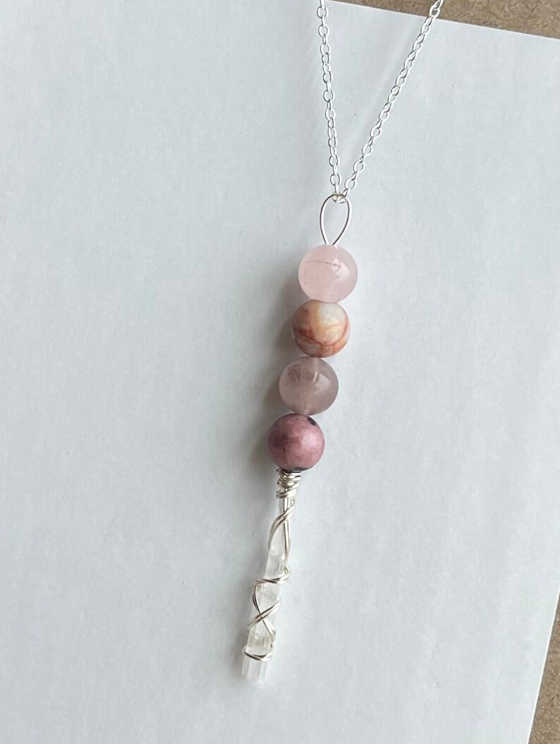May include: A silver chain necklace with a crystal point pendant wrapped in silver wire. The pendant is adorned with four pink and brown gemstone beads.
