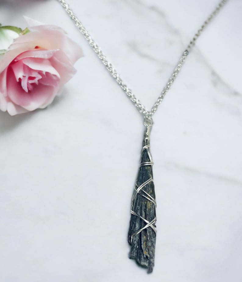 May include: A silver chain necklace with a dark gray, elongated pendant wrapped in silver wire. The pendant has a rough, textured surface. A pink rose is in the upper left corner of the image.