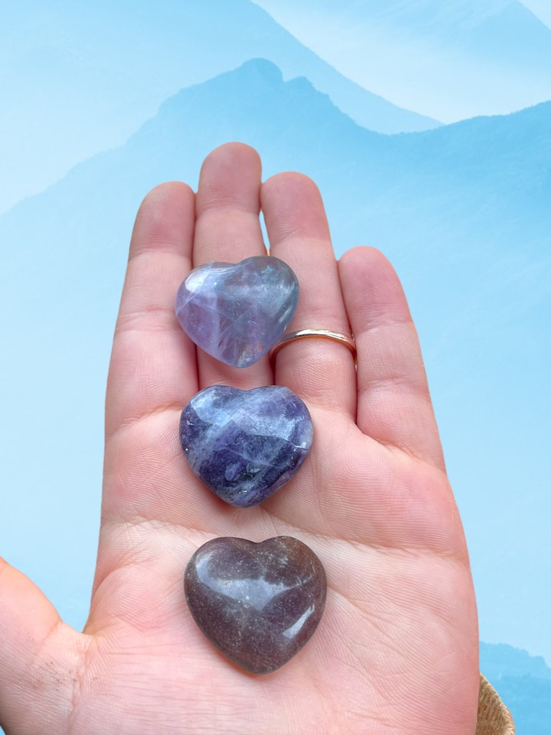 May include: Three heart-shaped amethyst crystals in shades of purple and lavender, held in a hand against a blue and white background.