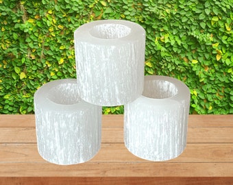 Può includere: Tre portacandele in selenite bianca, impilati su una superficie di legno. I portacandele sono cilindrici e hanno una superficie ruvida e testurizzata.