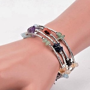 Op de afbeelding: Een meerstrengs wikkelarmband met zilverkleurig metaaldraad en kleine, gepolijste, onregelmatige edelsteensplinters in diverse kleuren zoals paars, groen, blauw en oranje, afgewisseld met kleine, ronde, zilverkleurige metalen kralen.