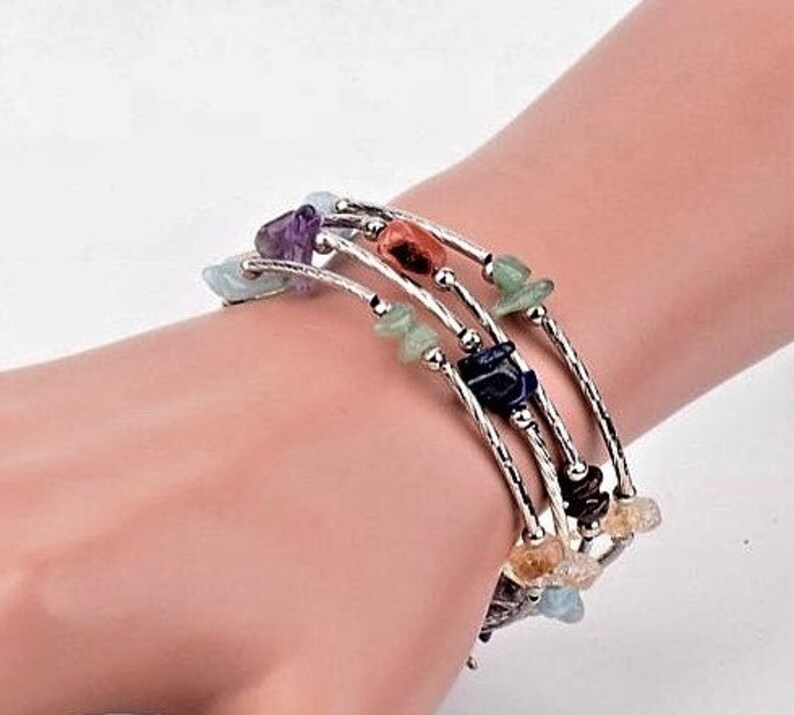 Op de afbeelding: Een meerstrengs wikkelarmband met zilverkleurig metaaldraad en kleine, gepolijste, onregelmatige edelsteensplinters in diverse kleuren zoals paars, groen, blauw en oranje, afgewisseld met kleine, ronde, zilverkleurige metalen kralen.