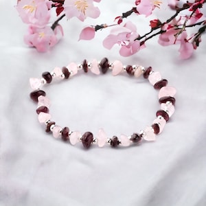 Op de afbeelding: Een armband met afwisselend rozenkwarts en granaatstenen, afgewisseld met kleine zilveren kralen. De armband wordt gepresenteerd op een witte achtergrond, met roze bloesem op de achtergrond.