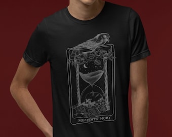 May include: A black t-shirt featuring a white graphic design of an hourglass with a bird perched on top. The graphic is surrounded by flowers and the text "Memento Mori" is printed below the hourglass.