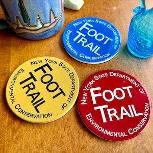 Puede incluir: Tres posavasos circulares con el logotipo del New York State Department of Environmental Conservation Foot Trail. Un posavasos es amarillo con texto negro, uno es azul con texto blanco y otro es rojo con texto blanco, todos circulares.