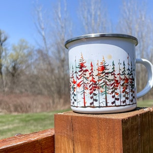 May include: White enamel camping mug with a colorful watercolor design of a forest scene. The mug has a stainless steel rim and handle.