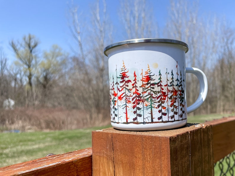 May include: White enamel camping mug with a colorful watercolor design of a forest scene. The mug has a stainless steel rim and handle.