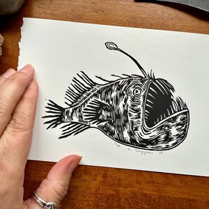 K&ouml;nnte beinhalten: Ein Schwarz-Wei&szlig;-Linolschnitt eines Anglerfischs. Der Druck zeigt die charakteristischen Merkmale des Fisches, darunter ein gro&szlig;es Maul, Flossen und einen biolumineszenten K&ouml;der. Die Signatur und das Datum des K&uuml;nstlers sind sichtbar.