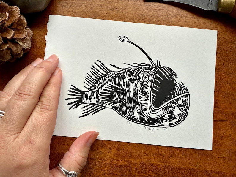 K&ouml;nnte beinhalten: Ein Schwarz-Wei&szlig;-Linolschnitt eines Anglerfischs. Der Druck zeigt die charakteristischen Merkmale des Fisches, darunter ein gro&szlig;es Maul, Flossen und einen biolumineszenten K&ouml;der. Die Signatur und das Datum des K&uuml;nstlers sind sichtbar.